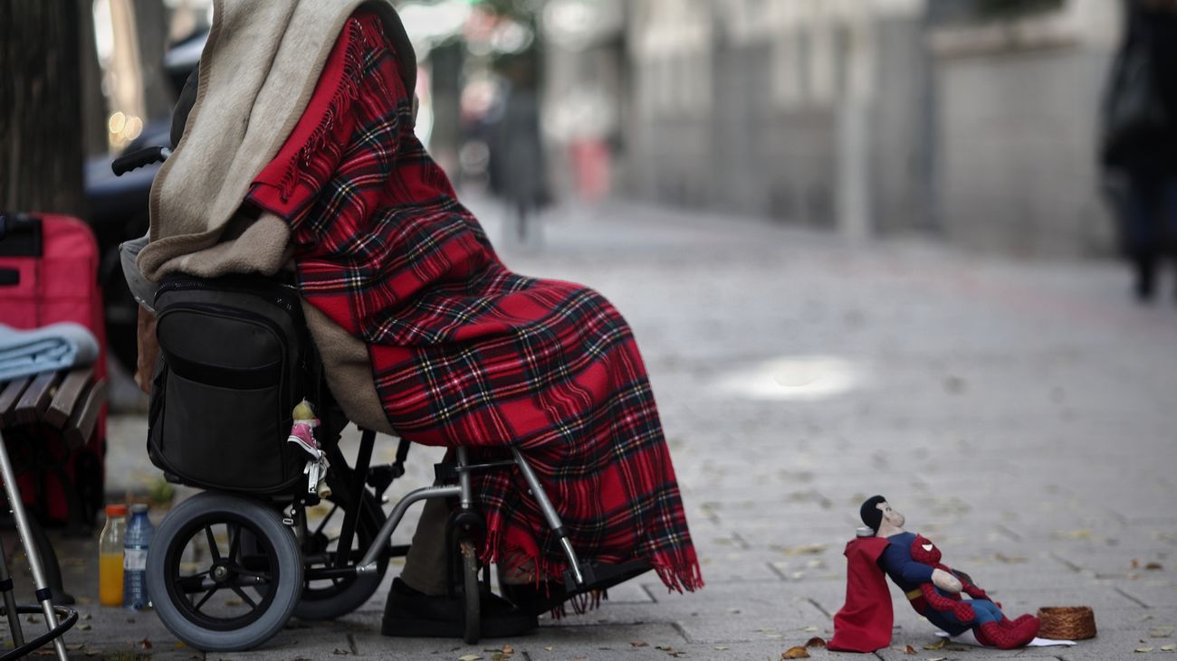 Una persona en silla de ruedas y tapada con una manta pide dinero con un muñeco de Superman en una céntrica calle