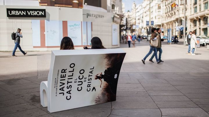 Dos personas se sientan en un banco con forma de libro de la campaña ‘Asienta la lectura-siéntate a leer’, en Gran Vía / EUROPA PRESS